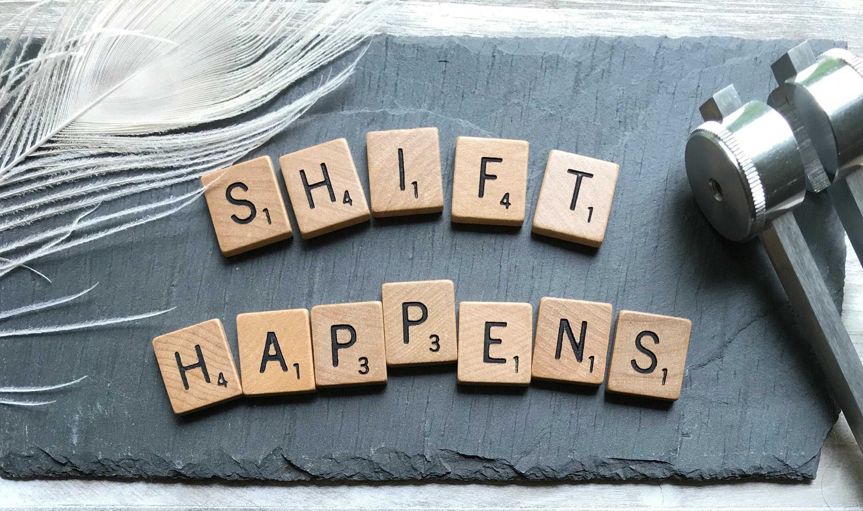 A picture of a slate with a feather and scrabble tiles spelling 'shift happens'